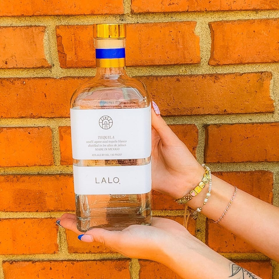 Hands holding a bottle of LALO tequila against a rustic brick wall background.