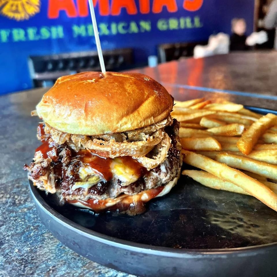 A close-up of a burger with pulled pork, onion rings, and fries on a dark plate.