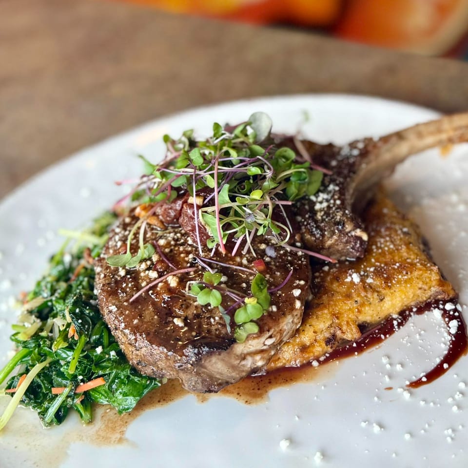 A bone-in pork chop with microgreens, served alongside greens and a golden-brown side dish.