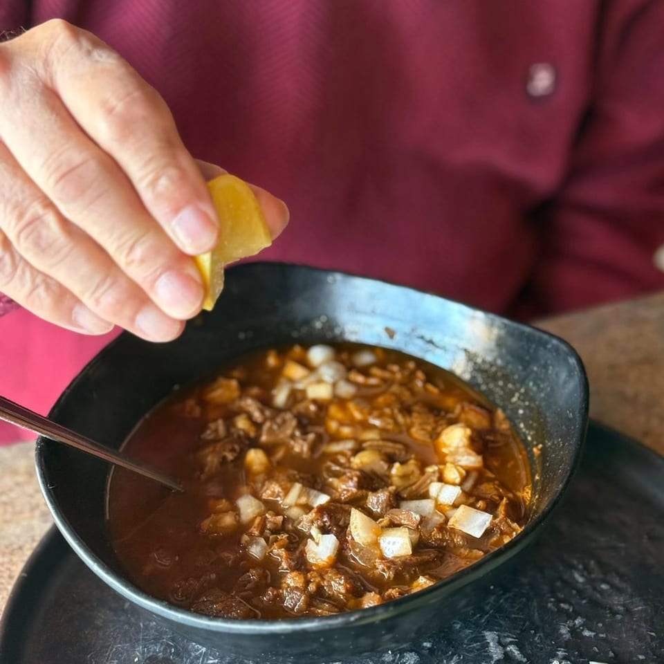 A hand squeezes a lemon wedge over a bowl of Amaya's Mexican beef stew with onions.