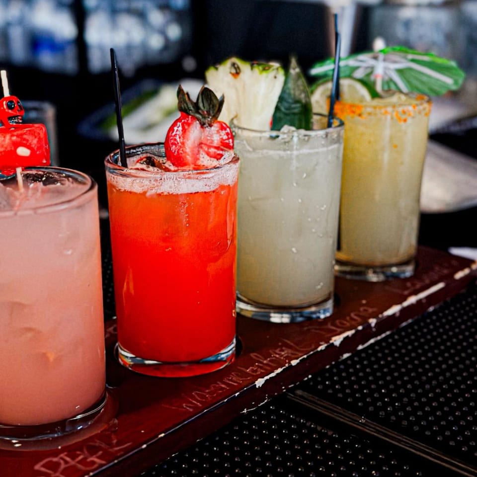 Four colorful Amaya's Fresh Mexican Grill cocktails, garnished with fruit and a tiny umbrella, on a wooden tray.