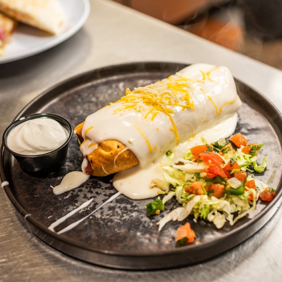 A large, smothered burrito with white sauce and shredded cheese, served with a side of sour cream and salad.