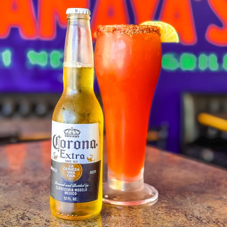 A Corona Extra beer bottle next to a red michelada cocktail with a lime wedge.