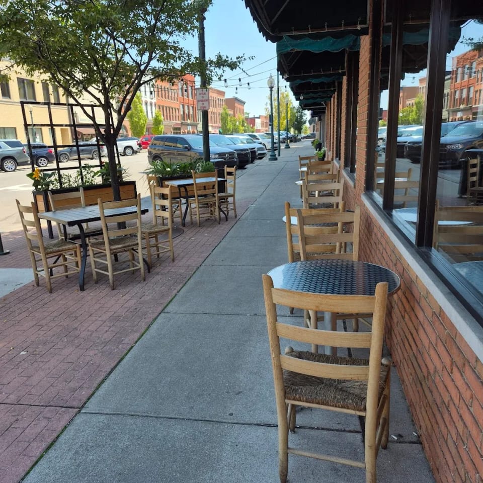 Outdoor patio seating with wooden tables and chairs lining a brick building on a city sidewalk.