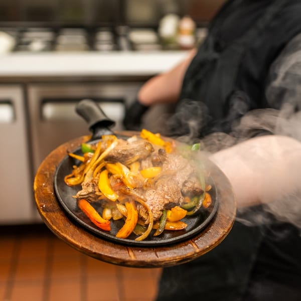 Sizzling beef fajitas with colorful peppers and onions on a hot skillet, held by a server.