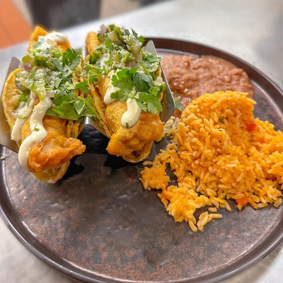Amaya's Fresh Mexican Grill's crispy tacos with cilantro, crema, rice, and refried beans on a dark plate.