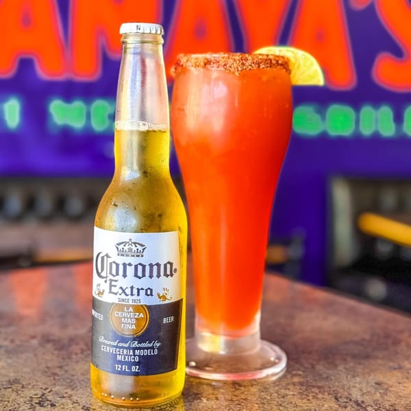 A Corona Extra beer bottle next to a tall glass of red michelada with a lime wedge.