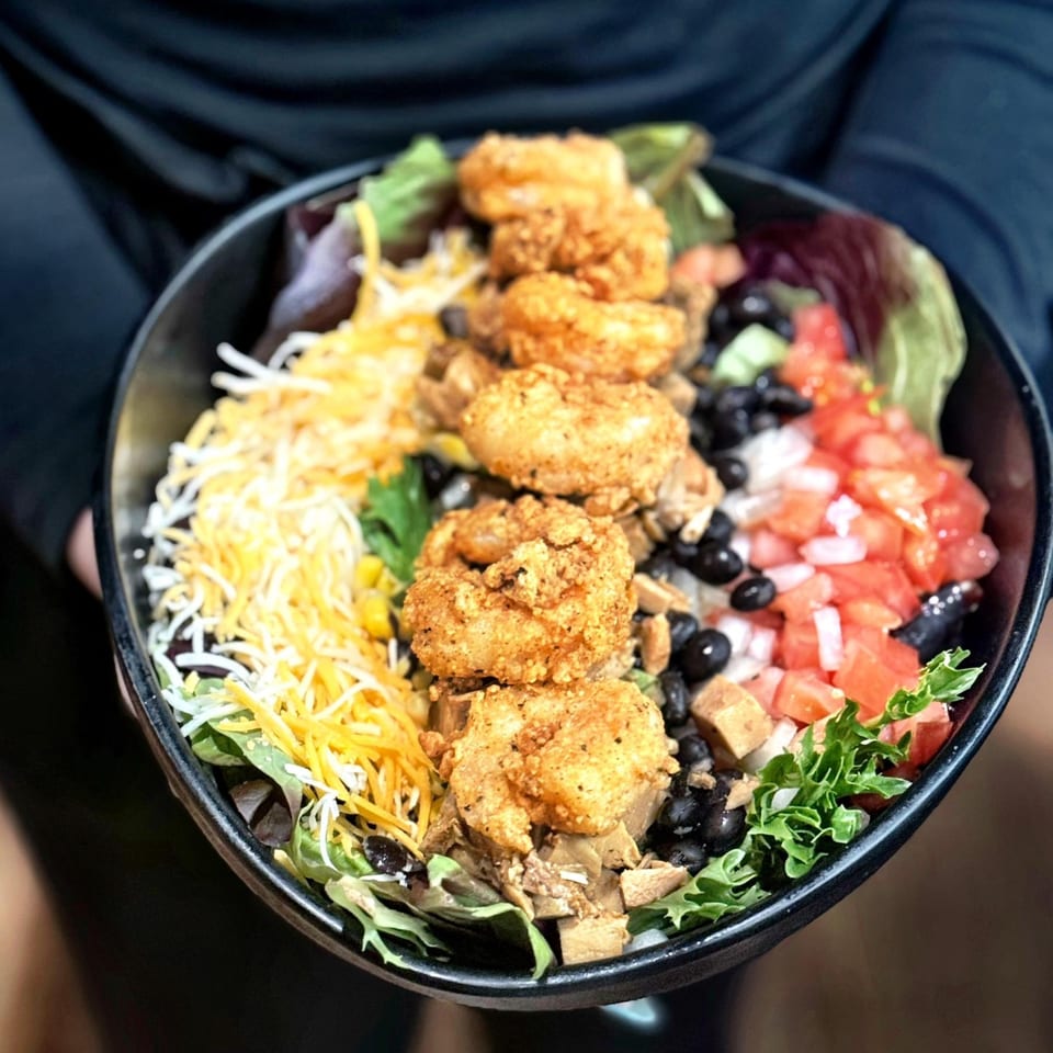 A person holds a large bowl of Amaya's Fresh Mexican Grill's shrimp salad with cheese, black beans, and tomatoes.