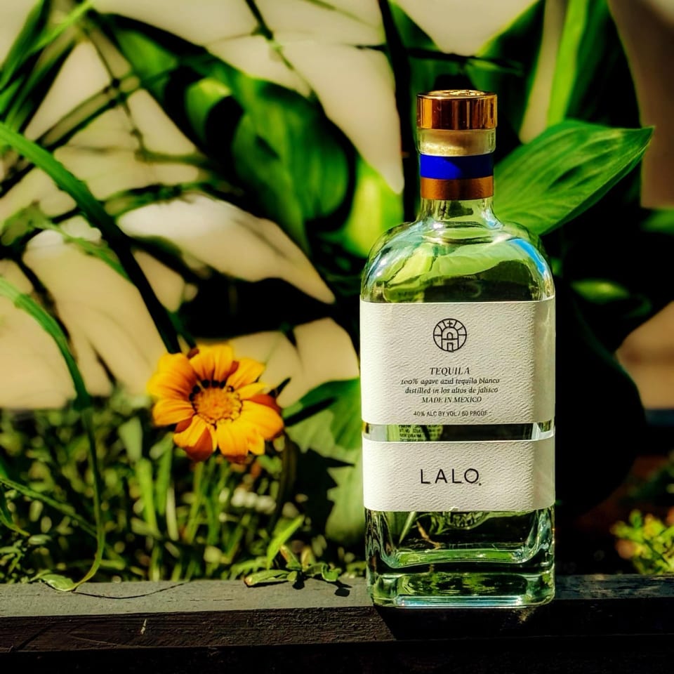 A bottle of LALO tequila with a gold cap and blue band, surrounded by green foliage and a yellow flower.