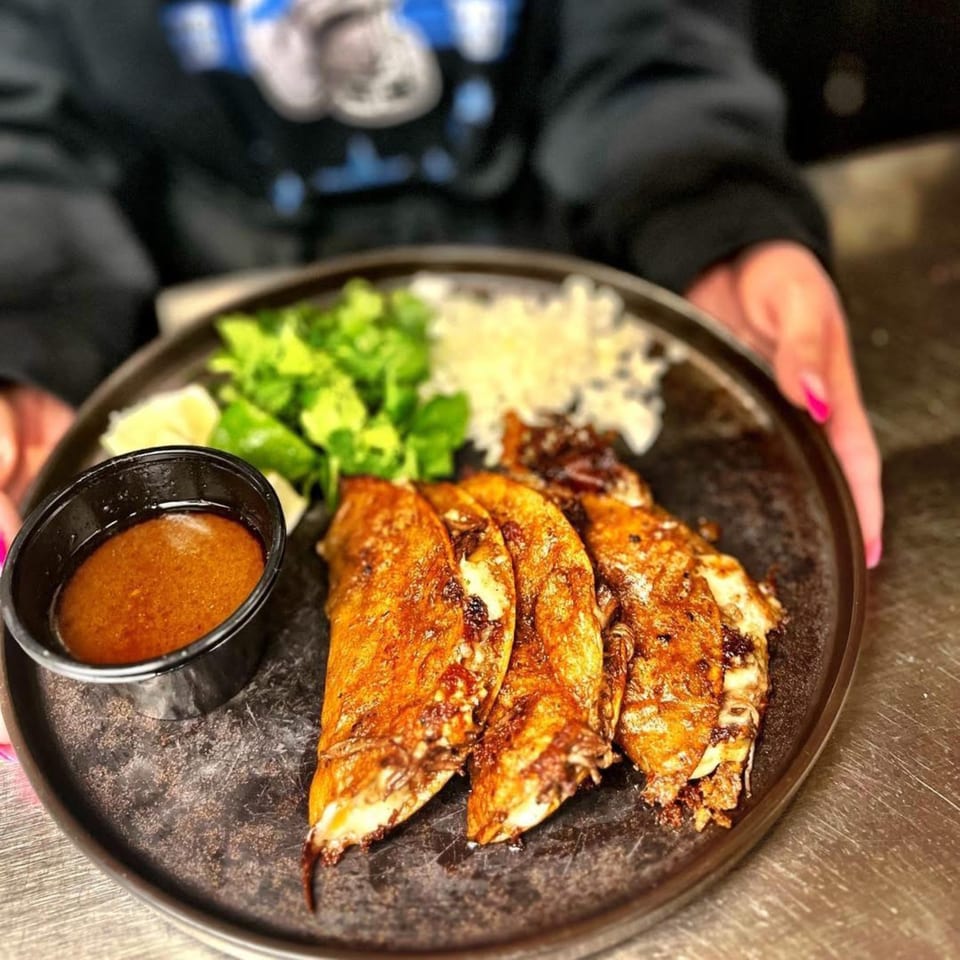 Hands holding a plate of Amaya's birria tacos with a side of cilantro, onions, lime, and dipping sauce.